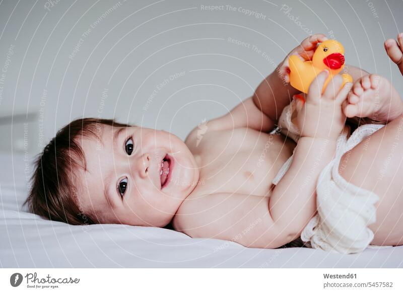 Lächelndes süßes Baby spielt mit Entenspielzeug, während es zu Hause auf dem Bett liegt Farbaufnahme Farbe Farbfoto Farbphoto Innenaufnahme Innenaufnahmen innen