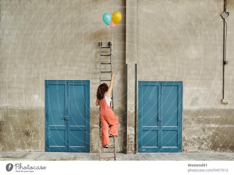 Frau klettert Leiter an Wand lehnend hinauf und greift nach bunten Heliumballons Farbaufnahme Farbe Farbfoto Farbphoto Innenaufnahme Innenaufnahmen innen