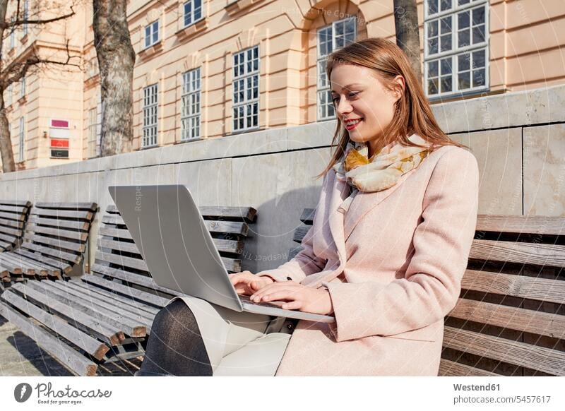 Österreich, Wien, lächelnde junge Frau sitzt auf Bank im MuseumsQuartier und benutzt Laptop weiblich Frauen benutzen benützen MQ sitzen sitzend Notebook Laptops