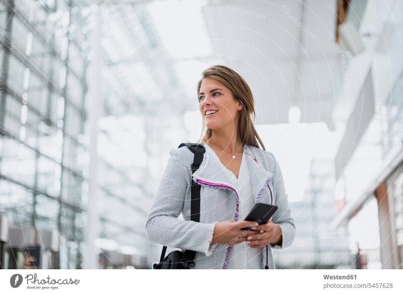 Porträt einer lächelnden jungen Geschäftsfrau mit Handy am Flughafen, die sich umsieht Mobiltelefon Handies Handys Mobiltelefone umherblicken umschauen umsehen
