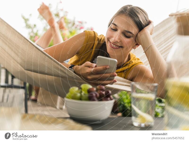 Lächelnde junge Frau benutzt Mobiltelefon, während sie sich auf Hängematte ausruht Farbaufnahme Farbe Farbfoto Farbphoto Deutschland Freizeitbeschäftigung Muße