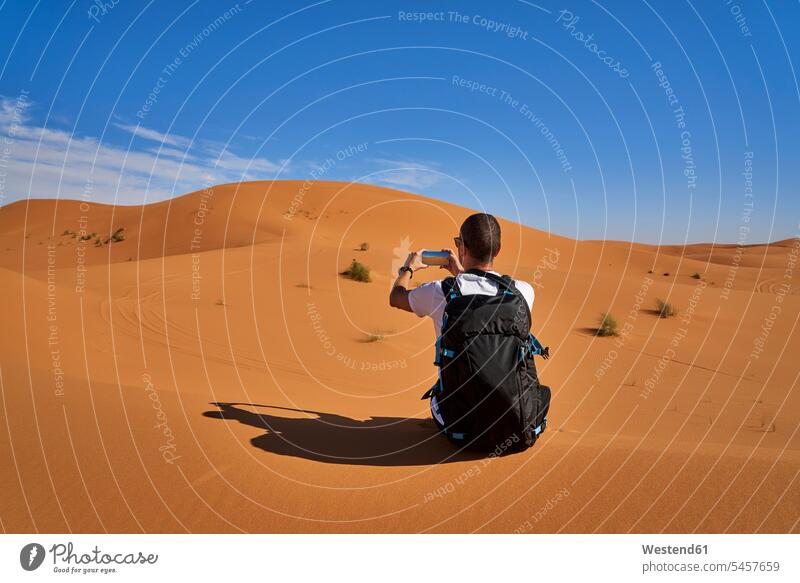 Marokko, Mann mit Rucksack sitzt auf Wüstendüne und fotografiert mit Smartphone iPhone Smartphones fotografieren sitzen sitzend Wüstendünen Wuestenduene