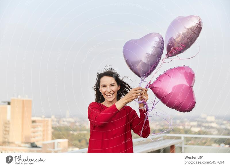 Lächelnde reife Frau hält herzförmigen Ballon in der Hand, während sie auf der Gebäudeterrasse steht Farbaufnahme Farbe Farbfoto Farbphoto Außenaufnahme außen