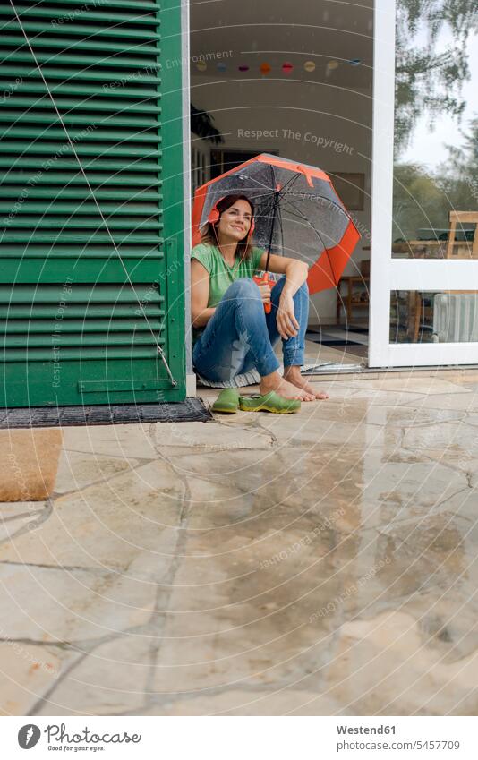 Lächelnde reife Frau sitzt auf der Terrasse im Regen unter Regenschirm und hört Musik mit Kopfhörern Schirme Regenschirme Kopfhoerer hoeren sitzend Terrassen