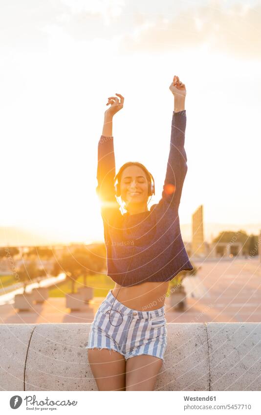 Spanien, Barcelona, Montjuic, glückliche junge Frau, die mit Kopfhörern bei Sonnenuntergang Musik hört Kopfhoerer hören hoeren Sonnenuntergänge Glück