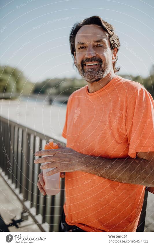 Porträt eines lächelnden Mannes, der eine Wasserflasche hält, während er im Park vor dem klaren Himmel steht Farbaufnahme Farbe Farbfoto Farbphoto Deutschland