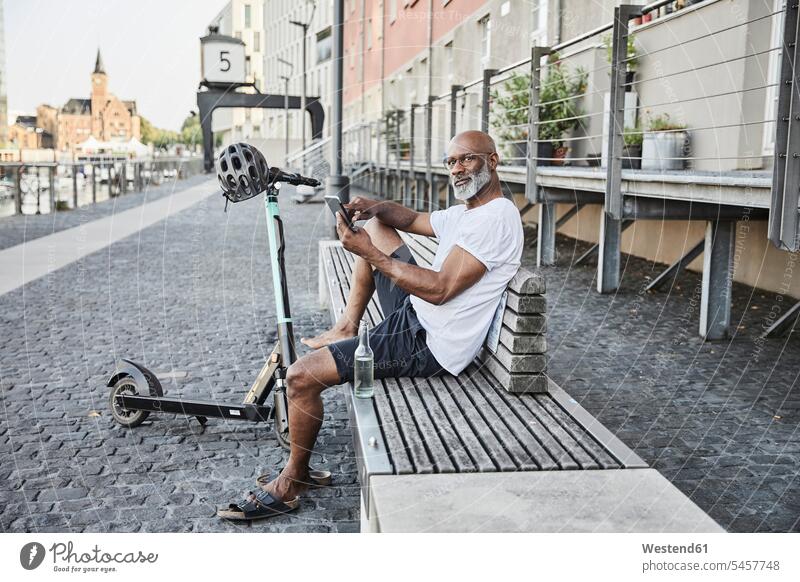 Porträt eines reifen Mannes mit E-Scooter, der sich im Sommer auf einer Bank ausruht und dabei sein Handy benutzt, Köln, Deutschland Flaschen Glasflaschen