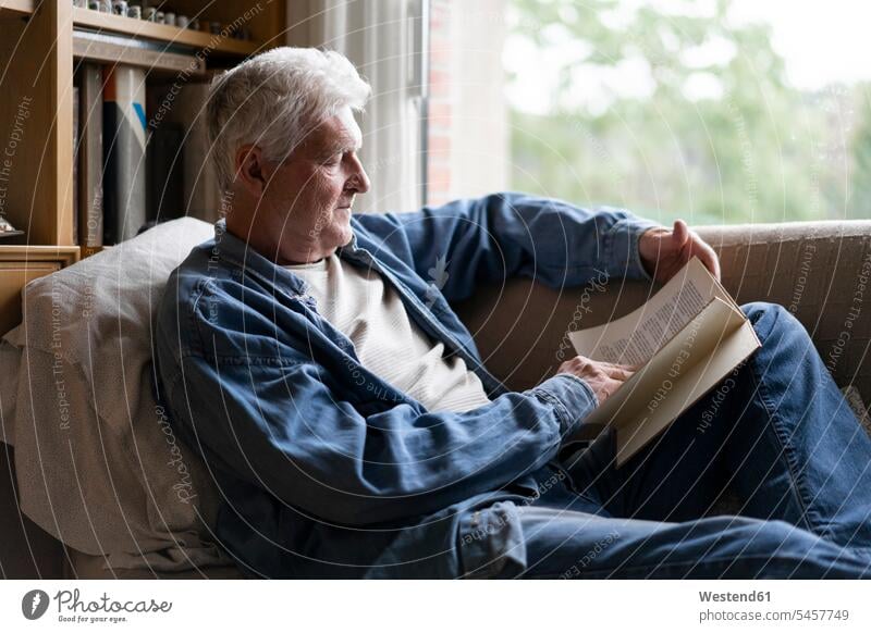 Älterer Mann liest Buch, während er sich zu Hause auf dem Sofa entspannt Farbaufnahme Farbe Farbfoto Farbphoto Innenaufnahme Innenaufnahmen innen drinnen Tag
