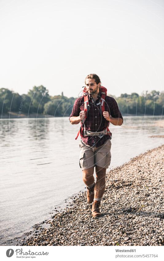 Junger Mann mit Rucksack beim Spaziergang am Flussufer Rucksäcke Fluesse Fluß Flüsse gehen gehend geht Männer männlich Gewässer Wasser Erwachsener erwachsen