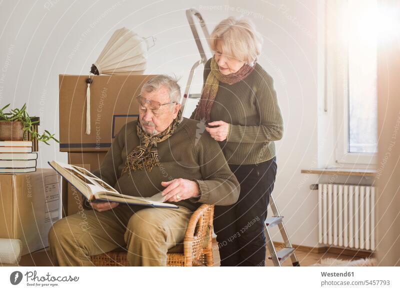 Älteres Ehepaar betrachtet Fotoalbum umgeben von Pappkartons in einem leeren Raum Kartons Kisten Bilder Bildnis Fotos Schals sitzend sitzt umziehen daheim