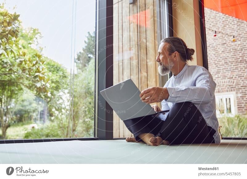 Bärtiger, reifer Mann mit Laptop, der durchs Fenster schaut, während er in einem winzigen Haus sitzt Farbaufnahme Farbe Farbfoto Farbphoto Deutschland