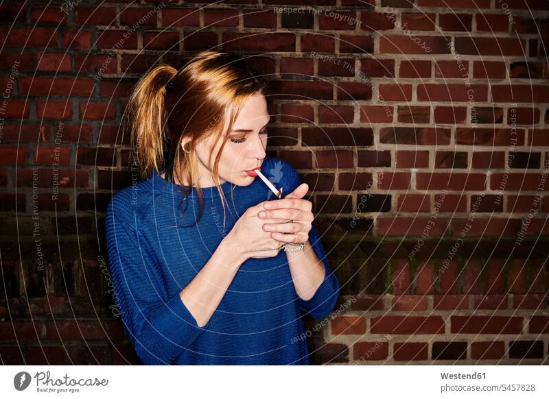 Junge Frau zündet nachts eine Zigarette gegen eine Ziegelmauer Farbaufnahme Farbe Farbfoto Farbphoto Außenaufnahme außen draußen im Freien Deutschland