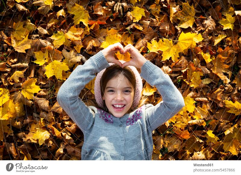 Porträt eines glücklichen kleinen Mädchens, das auf Herbstblättern liegt und mit ihren Händen ein Herz formt Portrait Porträts Portraits formen Herzform