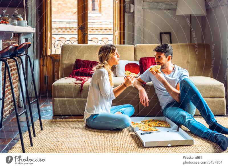 Junges Paar sitzt auf dem Boden des Wohnzimmers und isst Pizza aus dem Karton Boxen Kiste Schachtel Couch Couches Liege Sofas Teppiche sitzend freuen geniessen