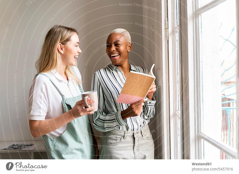 Zwei glückliche Frauen mit Notebook und Tasse Kaffee am Fenster Job Berufe Berufstätigkeit Beschäftigung Jobs geschäftlich Geschäftsleben Geschäftswelt