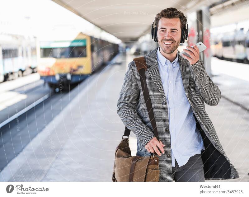 Lächelnder junger Geschäftsmann mit Handy und Kopfhörern am Bahnhof Leute Menschen People Person Personen Europäisch Kaukasier kaukasisch 1 Ein ein Mensch eine