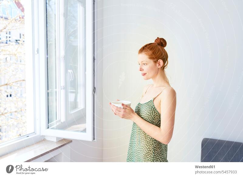 Porträt einer nachdenklichen Frau mit Schale mit weißem Kaffee am offenen Fenster stehend weiblich Frauen steht Schüssel Schalen Schälchen Schüsseln Portrait