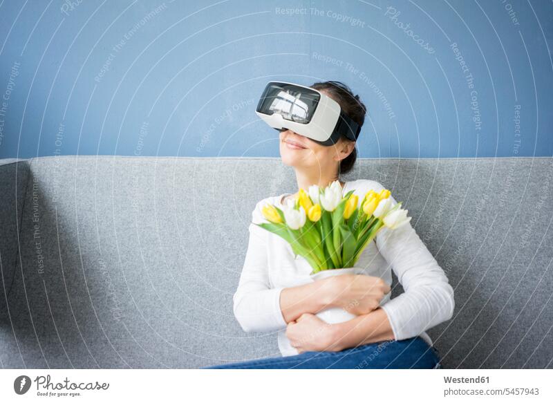 Lächelnde Frau sitzt auf Couch mit VR-Brille und hält Tulpenbüschel weiblich Frauen Blumenstrauß Bouquet Blumenstrauss Blumensträusse Blumensträuße sitzen