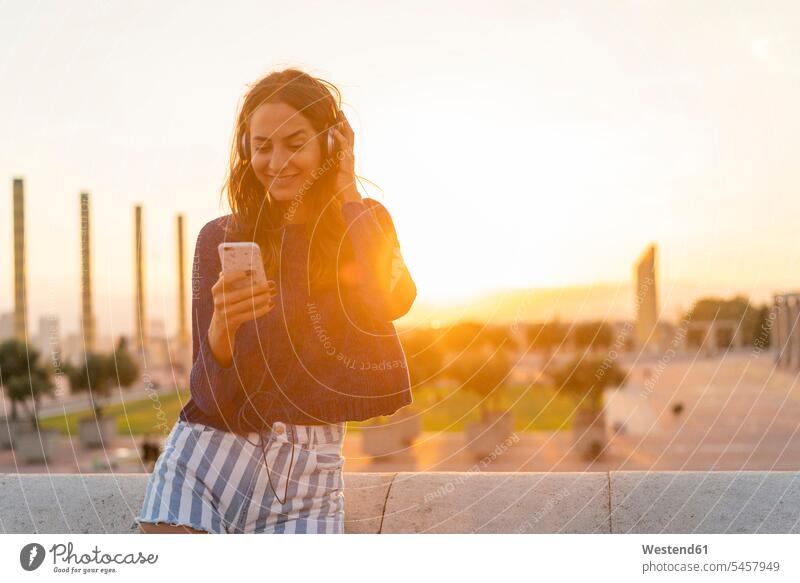 Spanien, Barcelona, Montjuic, lächelnde junge Frau mit Handy und Kopfhörer bei Sonnenuntergang Mobiltelefon Handies Handys Mobiltelefone Sonnenuntergänge