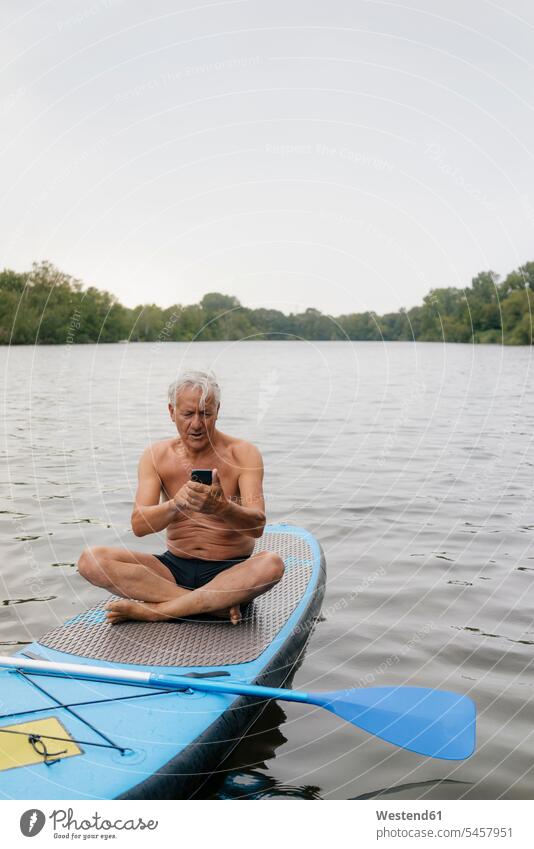 Senior Mann sitzt auf SUP-Board auf einem See mit Handy Männer männlich Seen Paddleboard Paddelbrett ältere Männer älterer Mann Senioren Mobiltelefon Handies