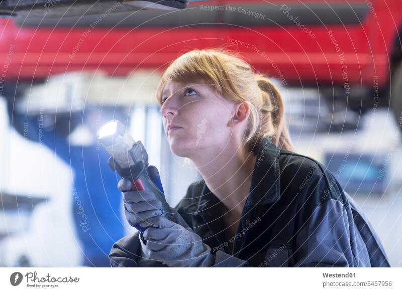 Automechanikerin mit Lampe, die auf den Boden eines Autos in der Reparaturwerkstatt schaut Mutige Frau Mutige Frauen Starke Frau Gewerbe Werkstaette
