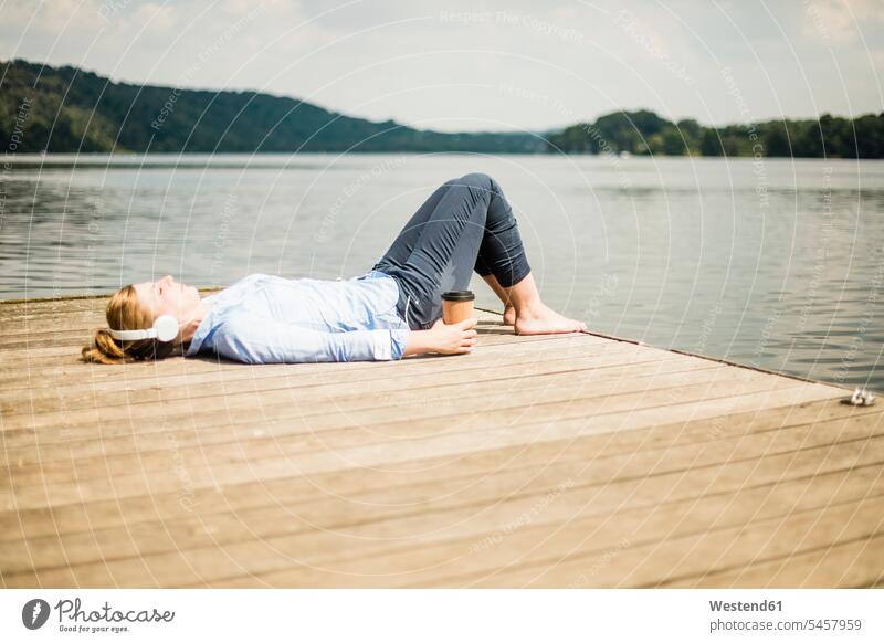 Frau liegend auf Steg an einem See mit Kopfhörer und Kaffee zum Mitnehmen Kopfhoerer Stege Anlegestelle liegt weiblich Frauen Seen Getränk Getraenk Getränke