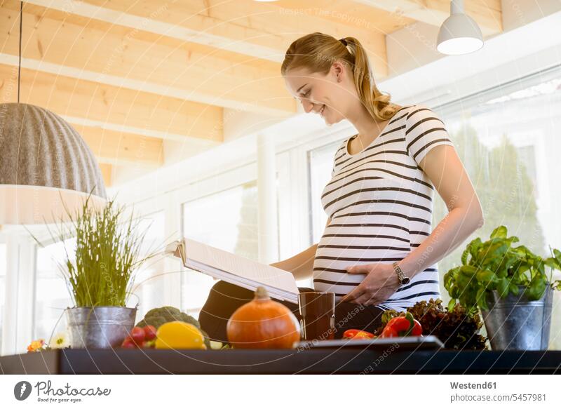 Lächelnde schwangere Frau liest zu Hause in der Küche Schwangere lesen Lektüre weiblich Frauen lächeln Buch Bücher Zuhause daheim Erwachsener erwachsen Mensch