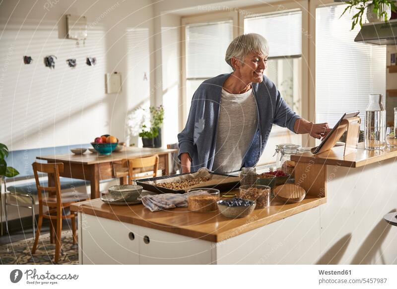 Lächelnde ältere Frau benutzt digitale Tablette in der Küche bei der Zubereitung von Müsli Leute Menschen People Person Personen Alleinstehende Alleinstehender