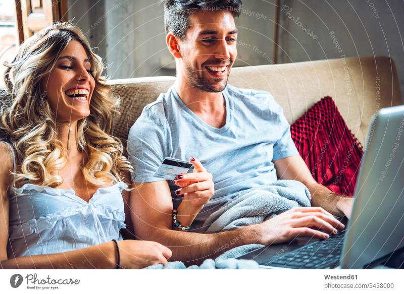Glückliches junges Paar sitzt auf Couch zu Hause mit Kreditkarte und Laptop Bankwesen EC-Karte Kreditkarten Karten Couches Liege Sofas Rechner Laptops Notebook