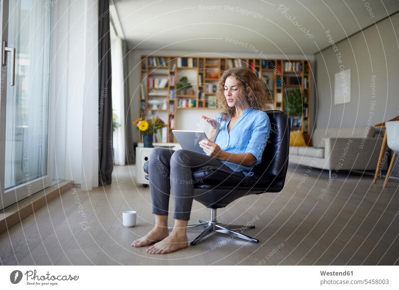 Frau bläst Kuss zu Videoanruf auf digitalem Tablet, während sie zu Hause auf einem Stuhl sitzt Farbaufnahme Farbe Farbfoto Farbphoto Innenaufnahme