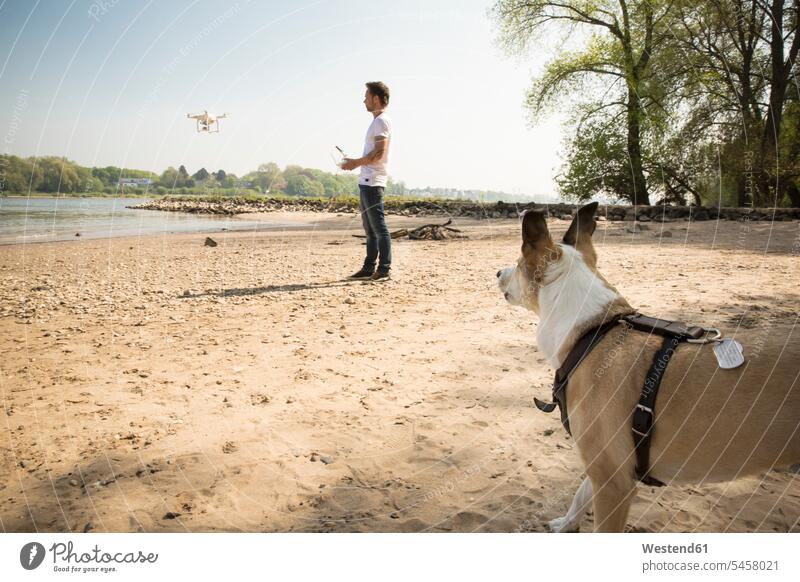 Mann mit Hund fliegt Drohne an einem Fluss Fluesse Fluß Flüsse Drohnen Männer männlich fliegen fliegend Hunde Gewässer Wasser Erwachsener erwachsen Mensch