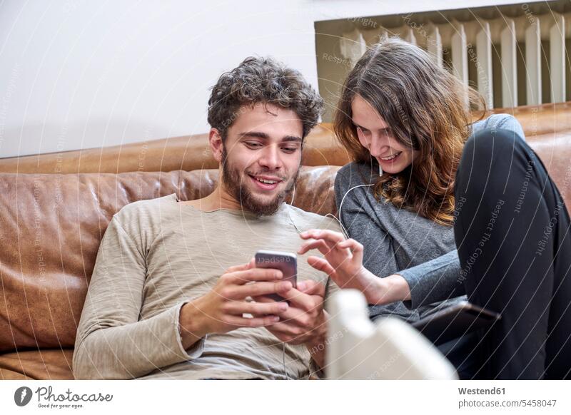Junger Mann und junge Frau sitzen auf der Couch und teilen sich Handy und Kopfhörer Mobiltelefon Handies Handys Mobiltelefone Teilen Sharing Paar Pärchen Paare