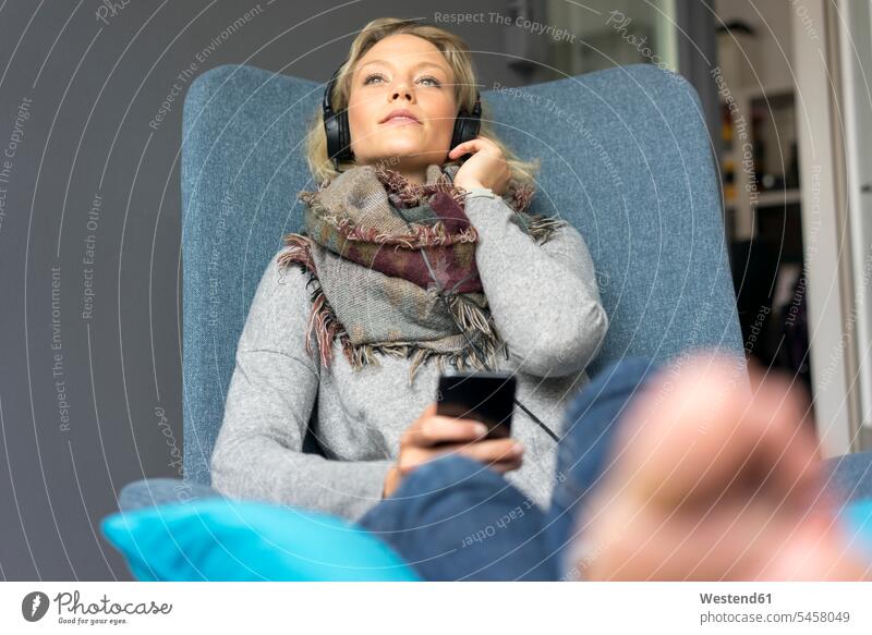 Junge Frau sitzt im Sessel und hört Musik mit Kopfhörern Schals Stuehle Stühle Kopfhoerer Telekommunikation telefonieren Handies Handys Mobiltelefon