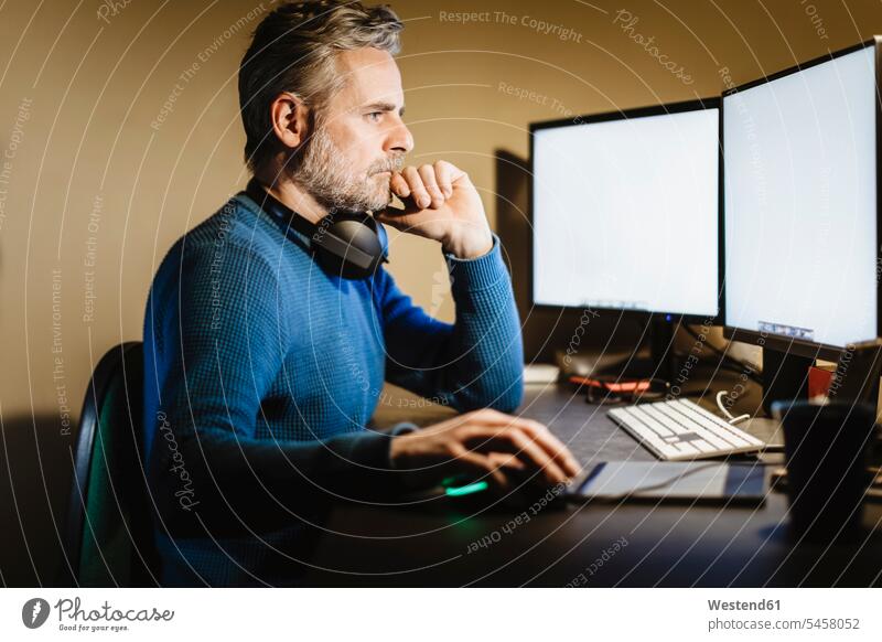 Reifer Mann sitzt zu Hause am Schreibtisch und arbeitet am Computer Job Berufe Berufstätigkeit Beschäftigung Jobs Tische Arbeitstisch Schreibtische Kopfhoerer