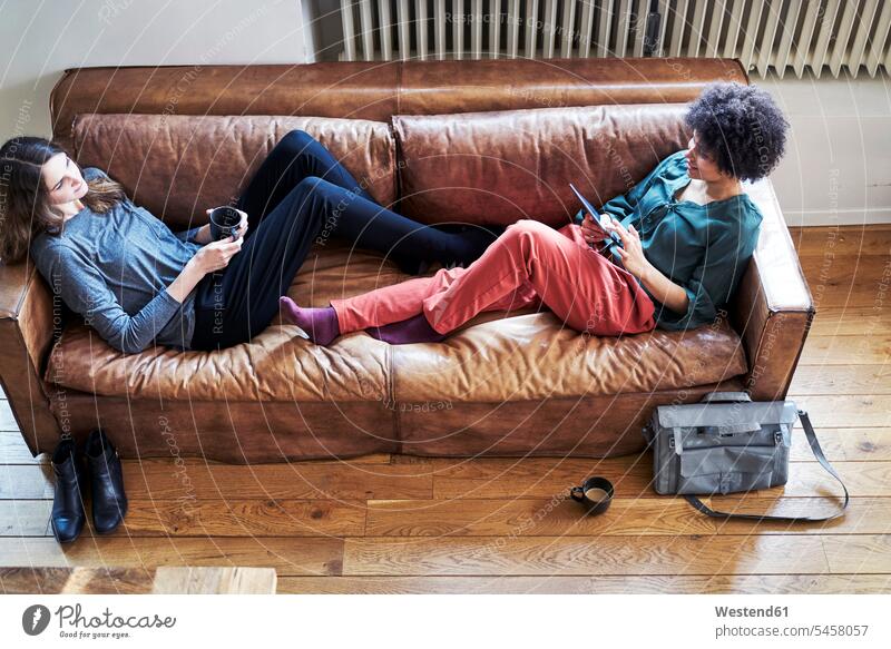 Zwei junge Frauen liegen auf einer Couch mit Tasse Kaffee und Tablette Kaffeetasse Kaffeetassen Tablet Computer Tablet-PC Tablet PC iPad Tablet-Computer liegend