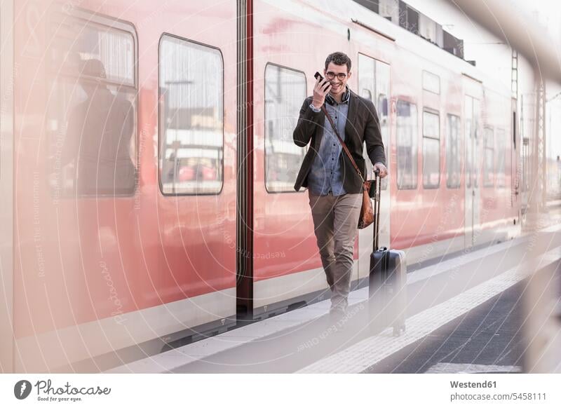 Glücklicher junger Mann mit Handy zu Fuß auf dem Bahnsteig entlang Nahverkehrszug gehen gehend geht Zugbahnsteig Zugbahnsteige Bahnsteige Bahnhof Bahnhöfe