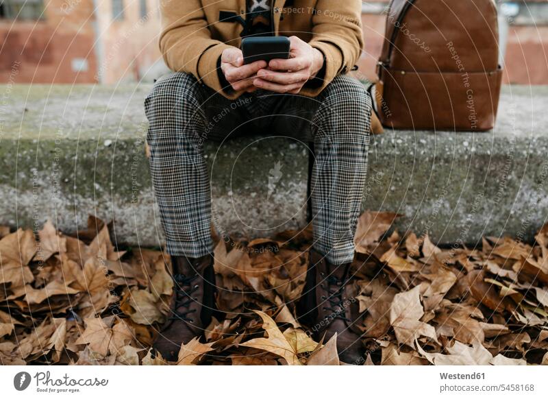 Spanien, Igualada, Nahaufnahme eines sitzenden Mannes, der ein Mobiltelefon benutzt, umgeben von Herbstblättern Männer männlich Herbstblatt Herbstblaetter sitzt