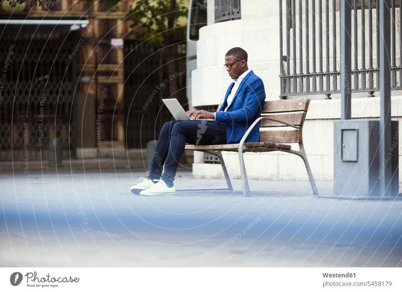 Junger Geschäftsmann in blauer Anzugjacke sitzt auf Bank und benutzt Laptop Leute Menschen People Person Personen Afrikanisch Afrikanische Abstammung