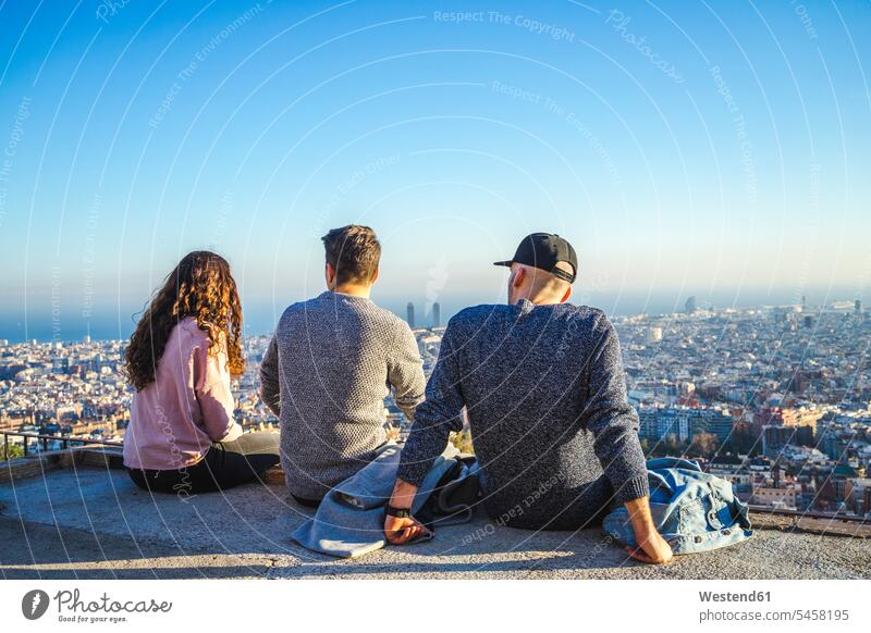 Spanien, Barcelona, drei Freunde sitzen auf einer Mauer mit Blick auf die Stadt sitzend sitzt überblicken staedtisch städtisch Mauern Außenaufnahme draußen
