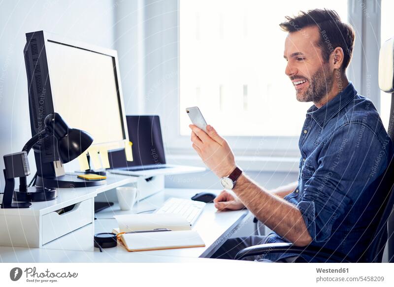Lächelnder Mann benutzt Handy am Schreibtisch im Büro Arbeitstisch Schreibtische Männer männlich Office Büros Mobiltelefon Handies Handys Mobiltelefone lächeln