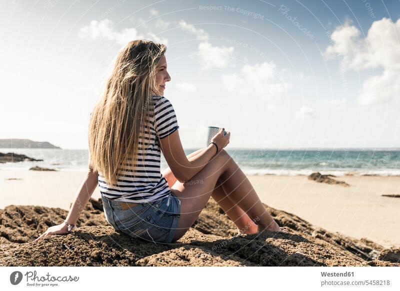Junge Frau sitzt in einem Felsen am Strand, entspannt mit einer Tasse Tee trinken sitzen sitzend Beach Straende Strände Beaches Pause Pause machen Entspannung