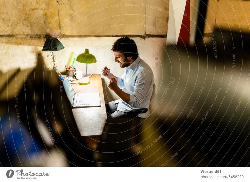 Jubelnder Geschäftsmann mit Laptop auf Tisch in der Dunkelheit Job Berufe Berufstätigkeit Beschäftigung Jobs geschäftlich Geschäftsleben Geschäftswelt