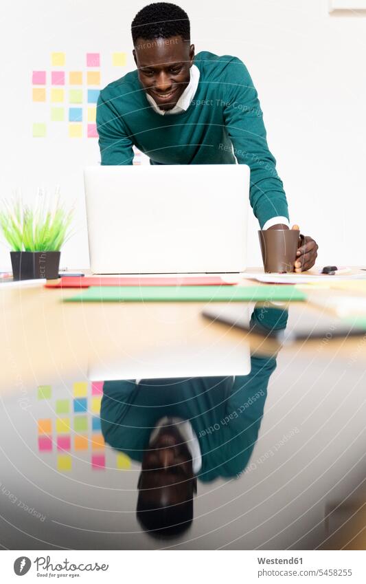 Lächelnder Mann arbeitet am Laptop am Schreibtisch im Büro Leute Menschen People Person Personen Afrikanisch Afrikanische Abstammung dunkelhäutig Farbige