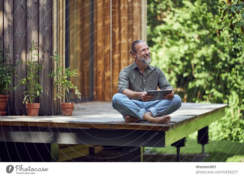 Bärtiger, reifer Mann hält digitales Tablett in der Hand und betrachtet, während er vor einem winzigen Haus sitzt Farbaufnahme Farbe Farbfoto Farbphoto