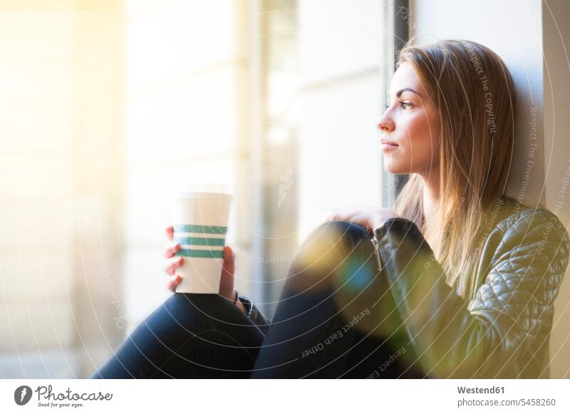 Frau schaut durch ein Fenster, während sie im Kaffeehaus eine Einweg-Kaffeetasse hält Farbaufnahme Farbe Farbfoto Farbphoto Innenaufnahme Innenaufnahmen innen