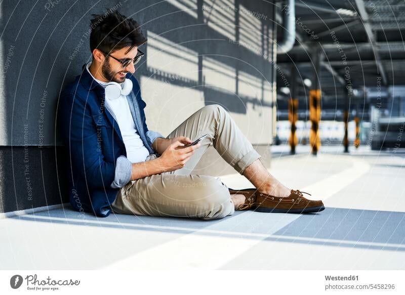 Lächelnder junger Mann, der auf dem Boden sitzt und ein Smartphone benutzt lächeln benutzen benützen iPhone Smartphones Männer männlich sitzen sitzend Handy