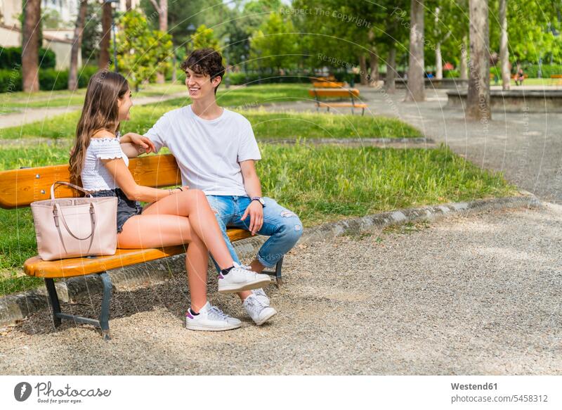 Junges Paar sitzt auf einer Bank und spricht miteinander in einem Park Taschen Handtaschen Bänke Sitzbank Sitzbänke sitzend reden sommerlich Sommerzeit freuen