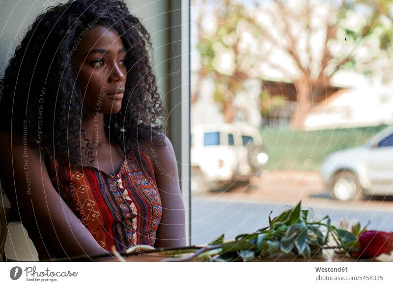 Porträt einer jungen afrikanischen Frau mit Blumen auf dem Tisch in einem Café, die aus dem Fenster schaut sitzend sitzt Erwartung sehnsüchtig Streben Muße