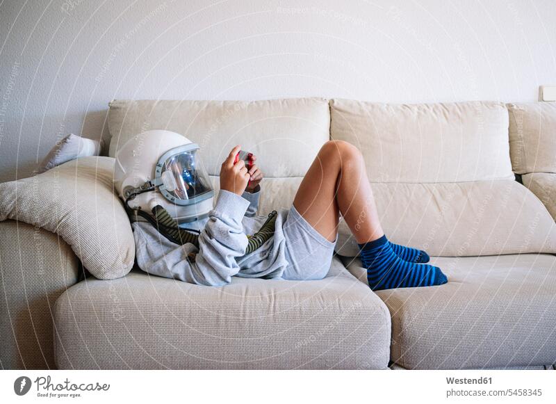 Junge mit Weltraumhelm spielt Videospiel, während er zu Hause auf dem Sofa liegt Farbaufnahme Farbe Farbfoto Farbphoto Spanien Innenaufnahme Innenaufnahmen