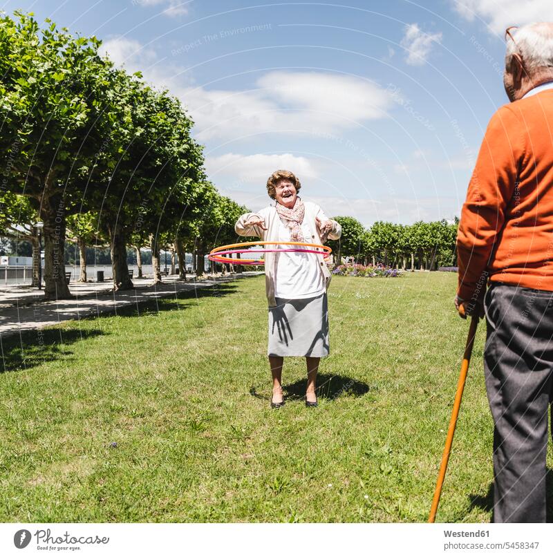 Senior beobachten ältere Dame spielen mit einem Hoola Hoop Park Parkanlagen Parks Sommer Sommerzeit sommerlich Seniorenpaar älteres Paar Seniorenpaare